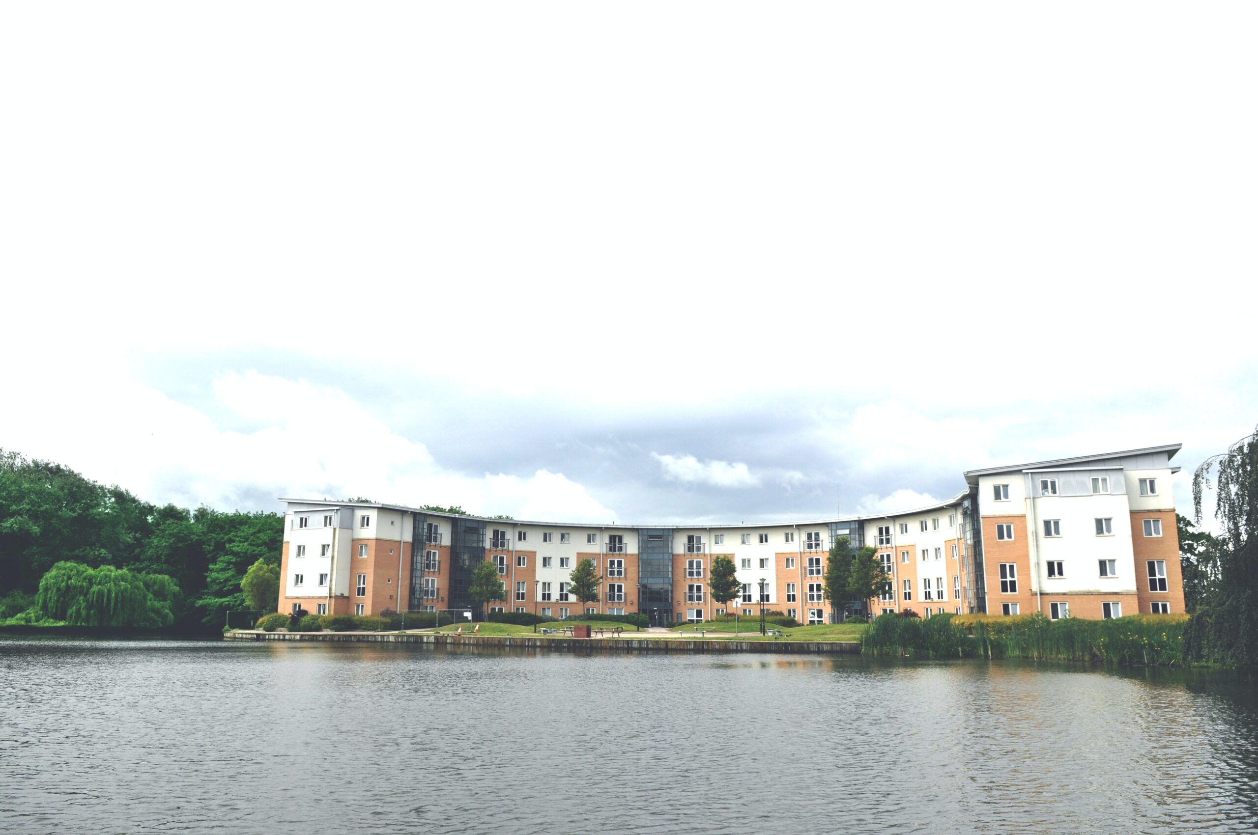 A large, curved apartment building with a mix of brick and white facade stands by a tranquil lake. The building is surrounded by greenery, with a cloudy sky overhead.