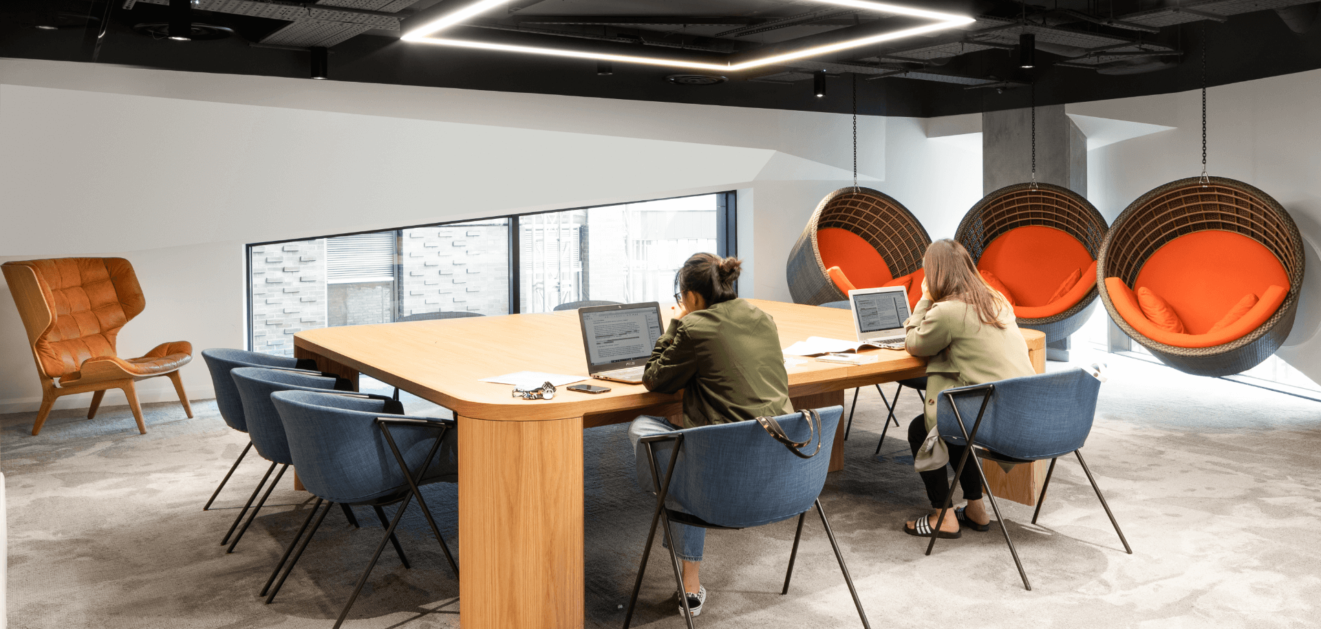 A modern office space with two people working on laptops at a wooden table. The room features hanging round chairs with orange cushions and a large window. The ceiling has a rectangular light fixture, and an orange chair is in the corner.