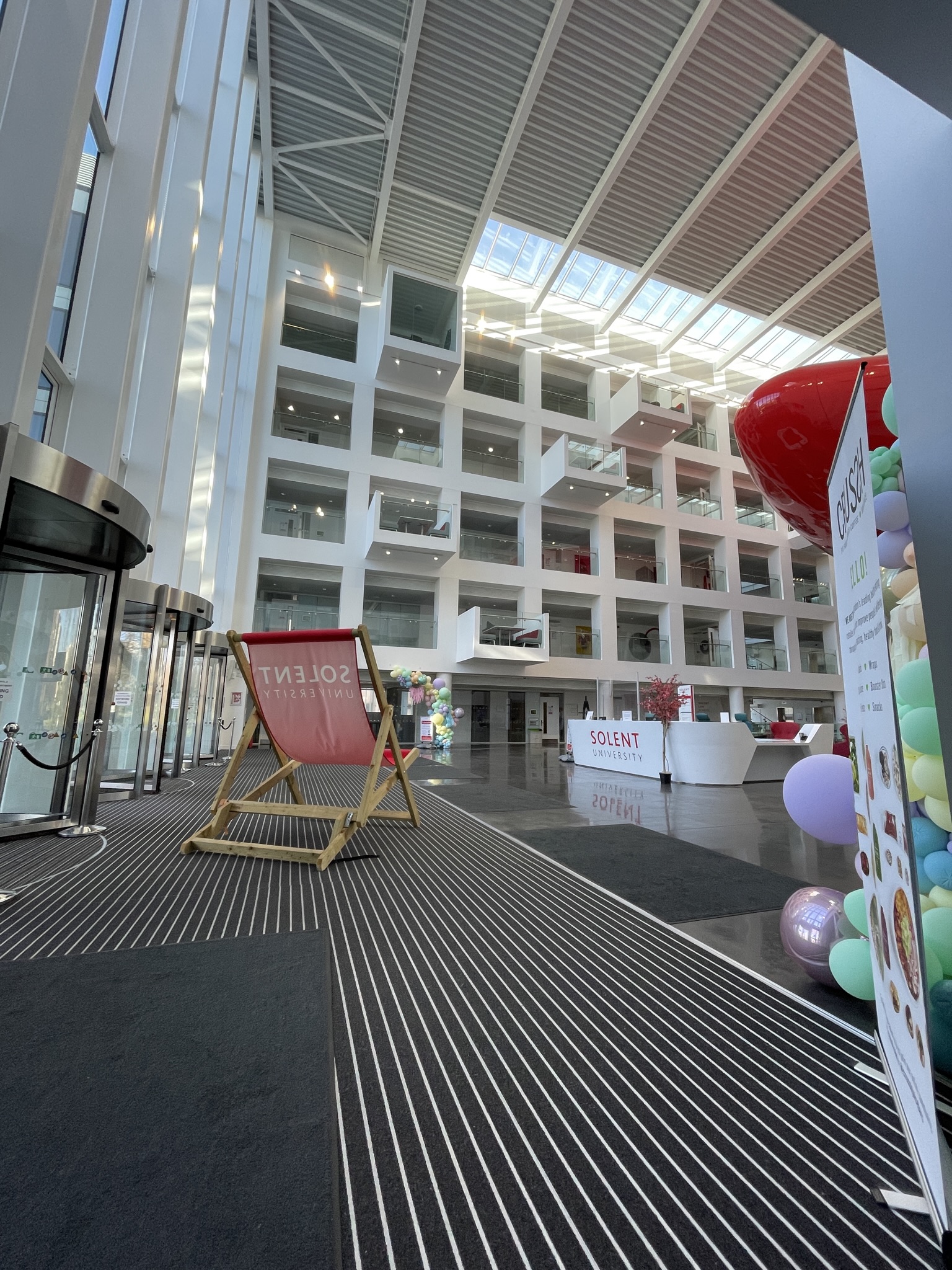 A modern, spacious building interior with high ceilings and large windows. The lobby features a seating area with a large deck chair and colorful balloons. A reception desk and glass entry doors are visible.
