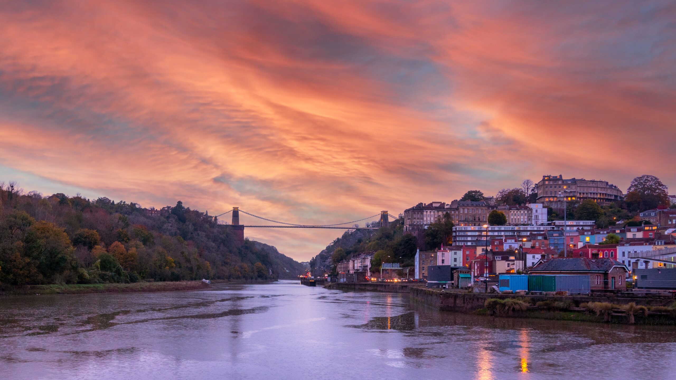 A picturesque view of a river at sunset with the Clifton Suspension Bridge in the background. The sky is vibrant with swirling pink and orange hues. Buildings line the riverbank to the right, while trees cover the left side.