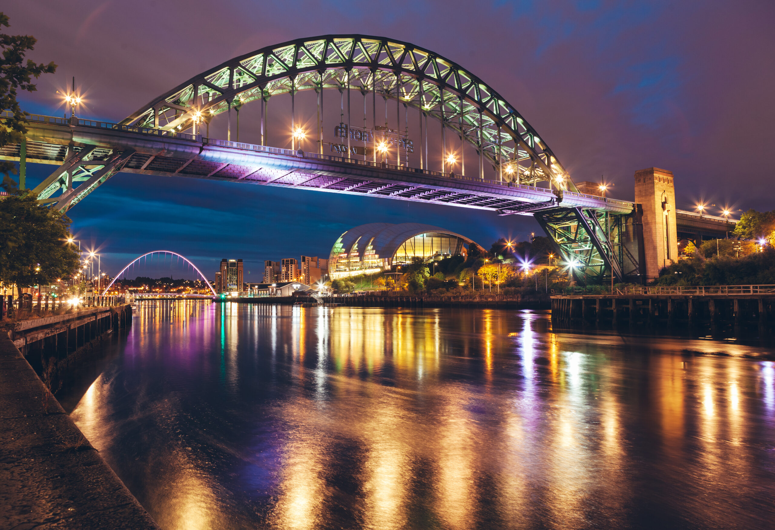 Night view of a lit-up bridge over a calm river, reflecting lights. The sky is cloudy, and city buildings are visible in the background, including a dome-shaped structure. The scene is serene with bright lights enhancing the bridge's structure.