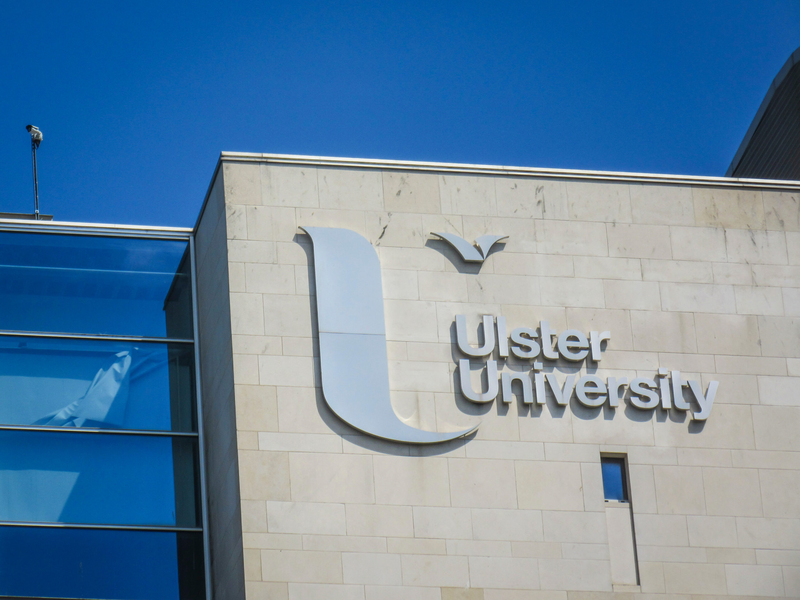 Exterior view of Ulster University with its name and logo displayed on a modern building facade under a clear blue sky.