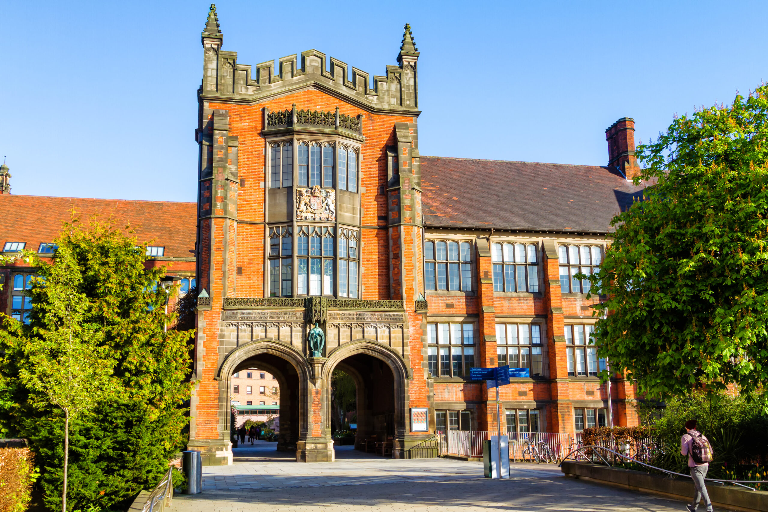 A large, historic red-brick building with an ornate central archway and decorative stonework. The architecture is framed by green trees, and a clear sky is visible. A few people walk nearby, and bicycles are parked to the right.