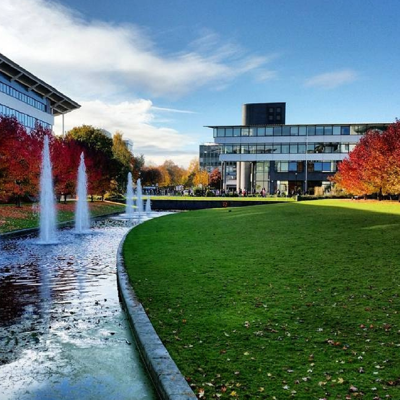 A modern student accommodation in Coventry is surrounded by vibrant autumn trees and green grass. Three fountains line a reflective water feature in the foreground, under a blue sky with wispy clouds.