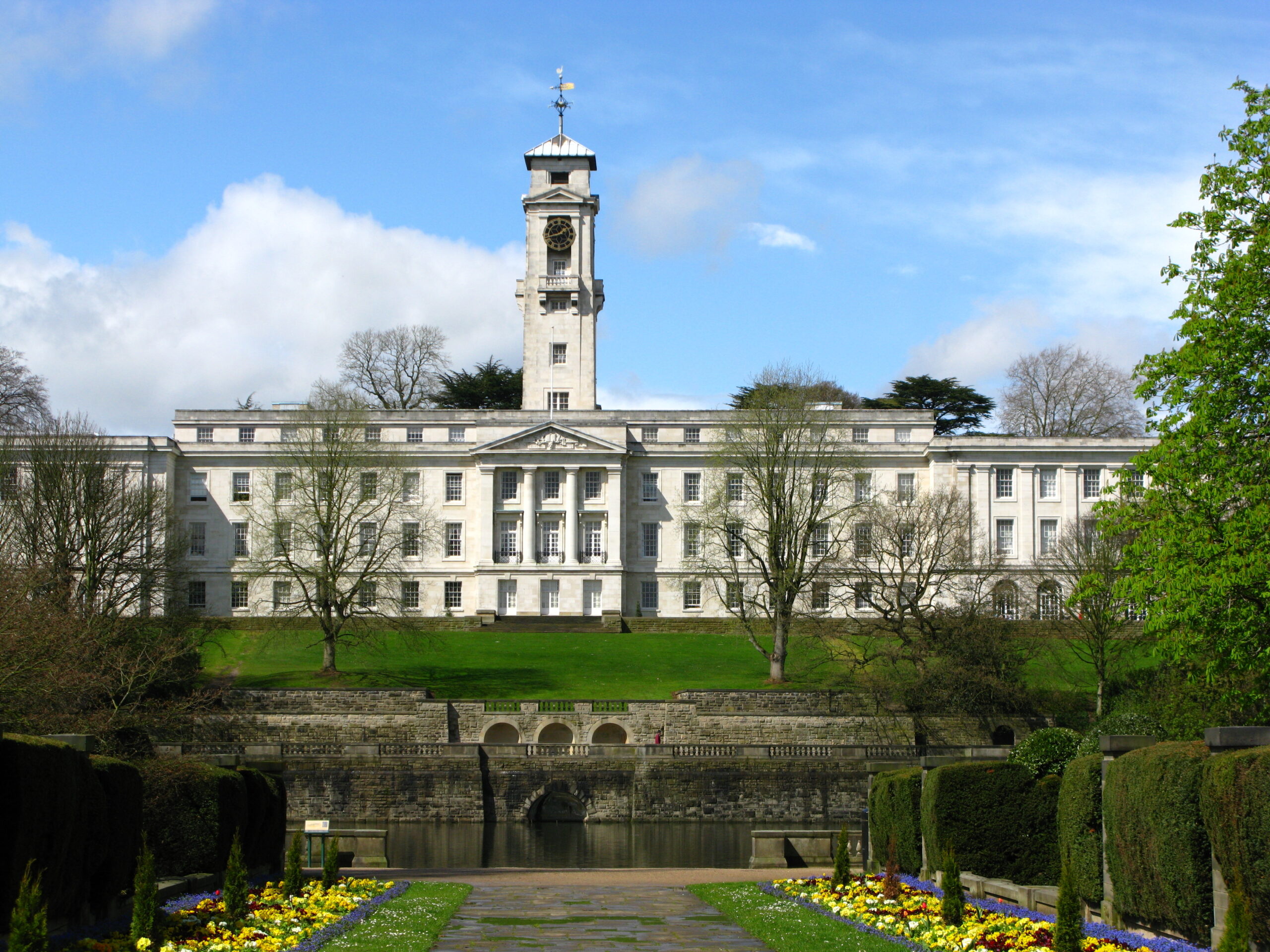 A grand white building with a tall clock tower stands amid green lawns and trees under a clear blue sky. A landscaped garden with colorful flowers and a stone path leads to the structure.