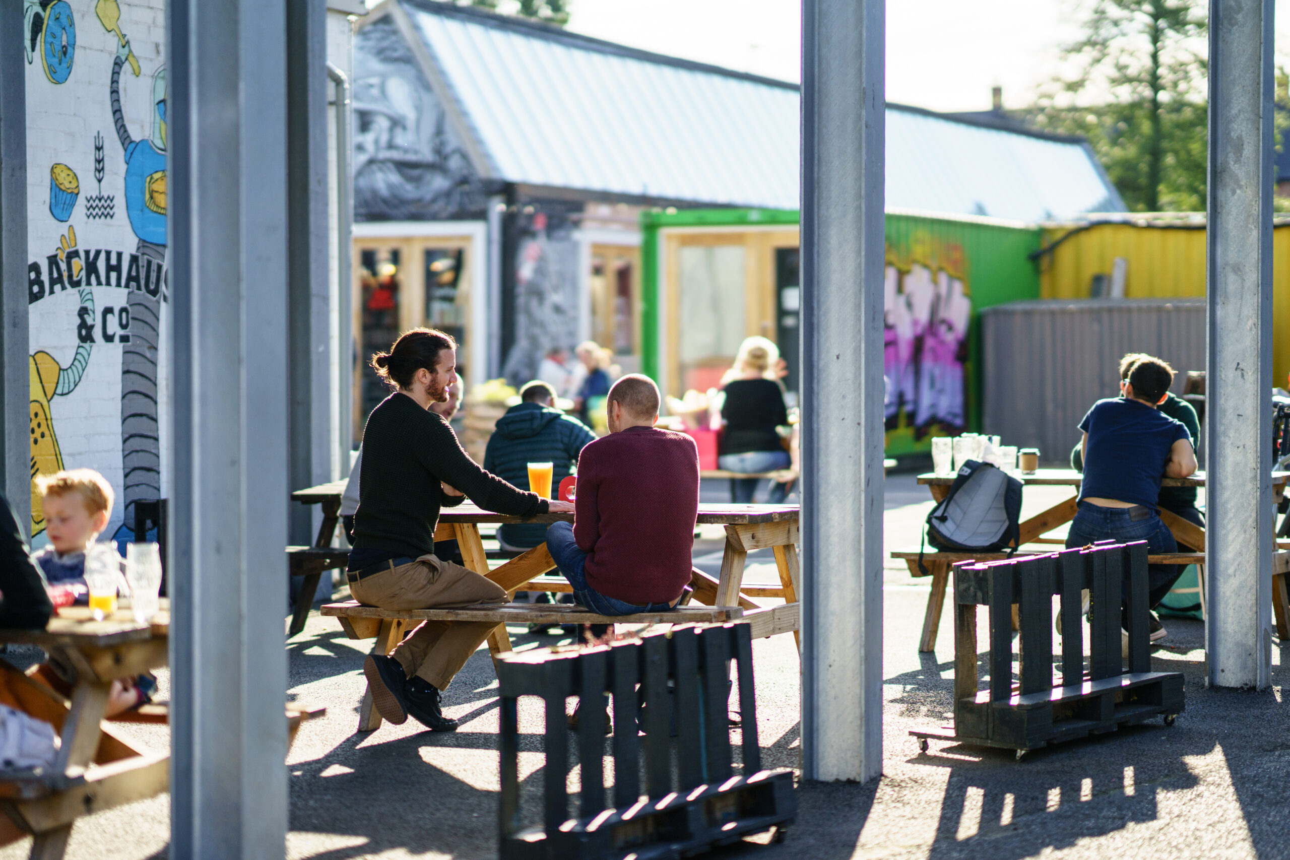 People sit at outdoor picnic tables, enjoying food and drinks in a casual setting near student accommodation in Coventry. Colorful murals adorn the surrounding buildings, creating a relaxed, open-air atmosphere with bright sunlight and patches of greenery.