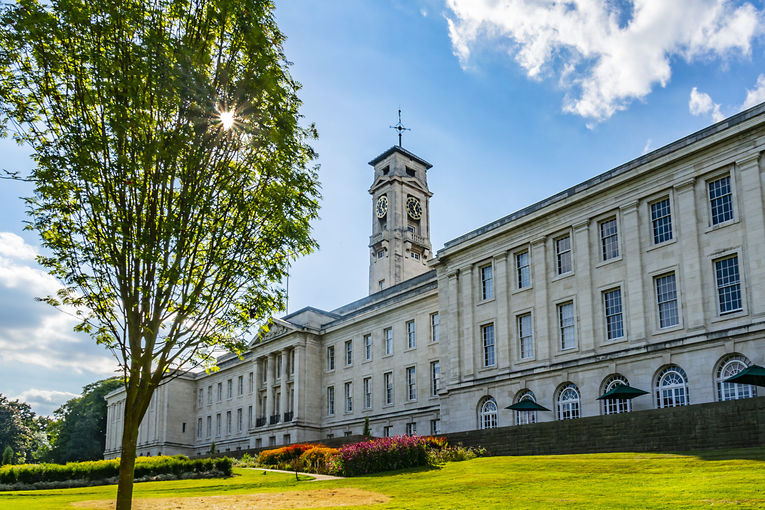 A large, historic building with a clock tower stands under a blue sky. Sunlight filters through the branches of a nearby tree, casting shadows on the lush green lawn and colorful garden in the foreground.