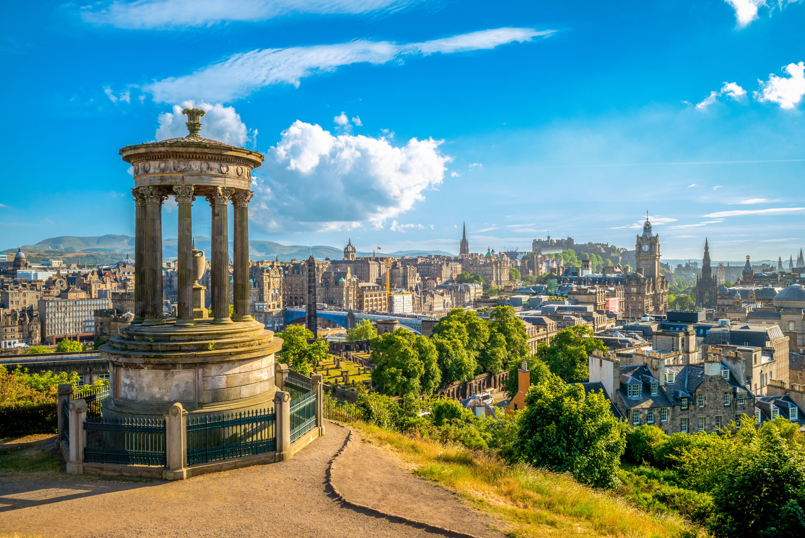Panoramic view of Edinburgh from Calton Hill, featuring the Dugald Stewart Monument in the foreground. The city's historic architecture, including Edinburgh Castle, is visible under a bright blue sky with scattered clouds.