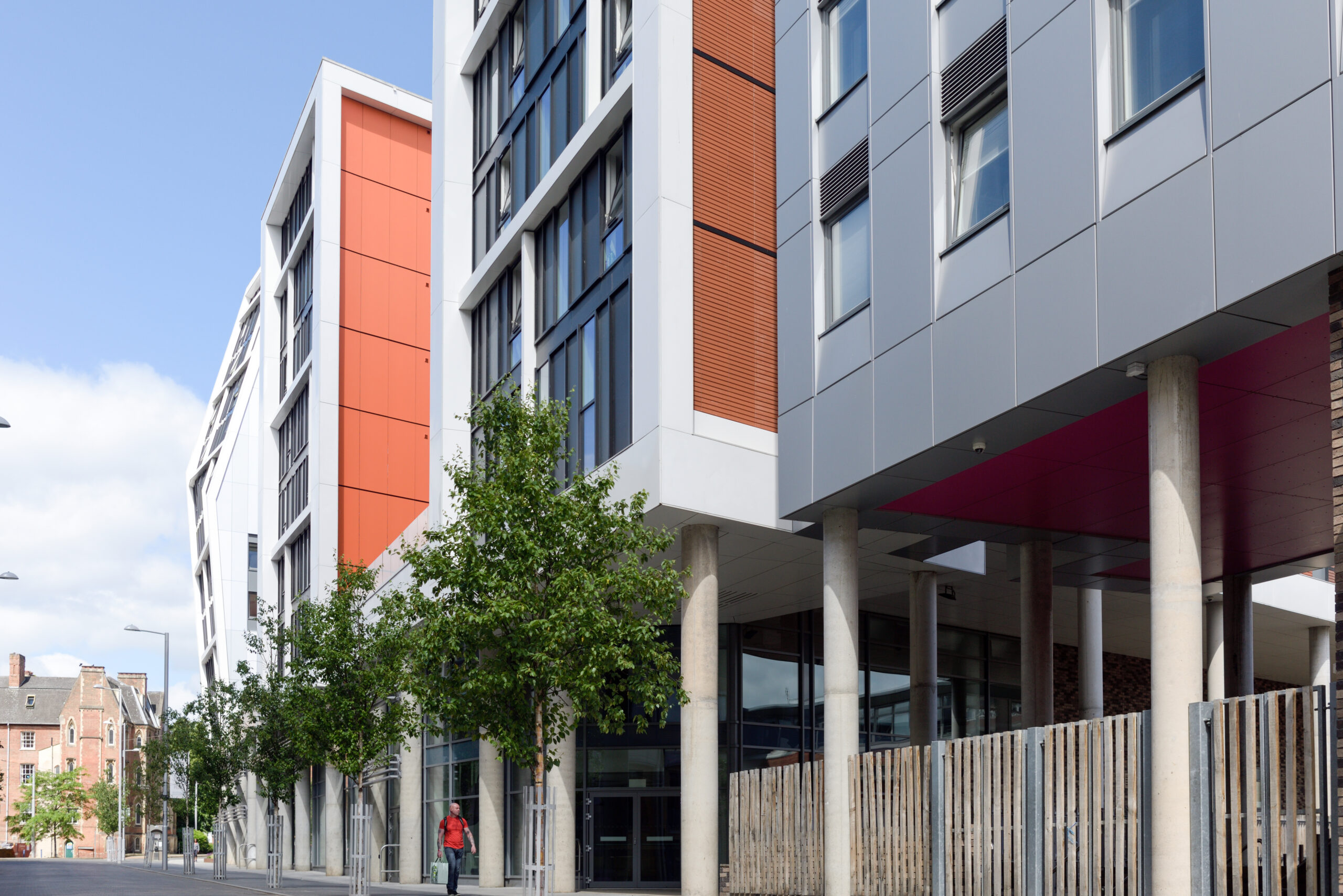 Modern building with tall glass windows and orange accents, supported by concrete pillars. Trees line the sidewalk, and a person in red walks along the street. The sky is partly cloudy.