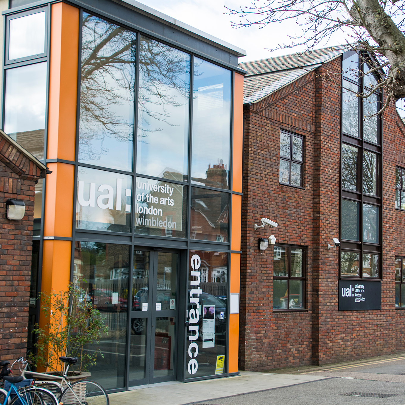 A modern brick building with large glass windows shows the entrance to the University of the Arts London, Wimbledon. The name and word "entrance" are visible on the glass. A bicycle is parked nearby, and bare trees are in the background.
