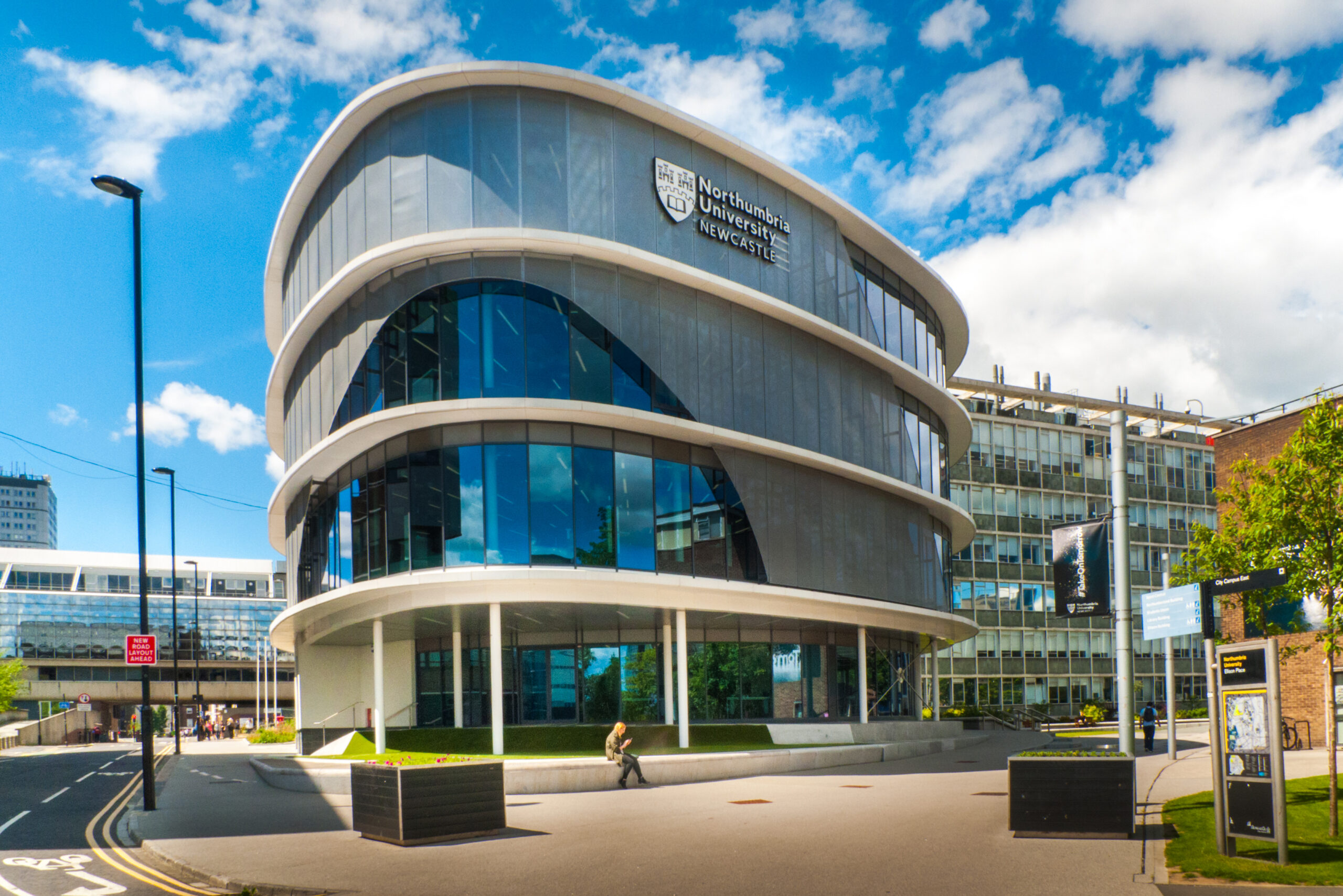 Modern, circular building with large glass windows, part of Northumbria University, Newcastle. The sleek design has multiple floors with a curved facade. The sky is partly cloudy and a few people are visible near the entrance.