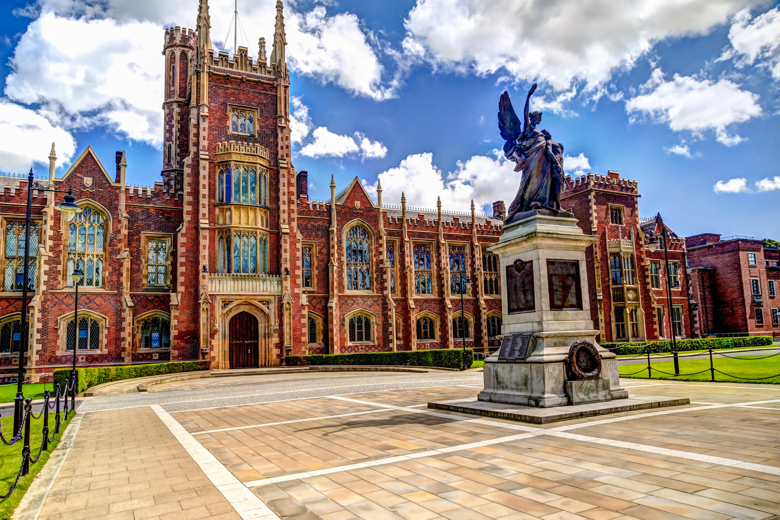 Historic Gothic-style building with ornate brick facade, tall arched windows, and a central tower, set against a partly cloudy sky. A large statue with an angelic figure stands prominently in the foreground on a paved plaza.