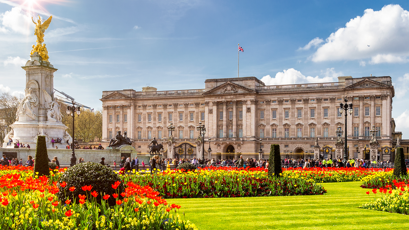 Buckingham Palace on a sunny day with a clear blue sky. The front is adorned with bright red and orange tulips in well-manicured gardens. The Victoria Memorial is in the foreground, and the Union Jack flies atop the palace.