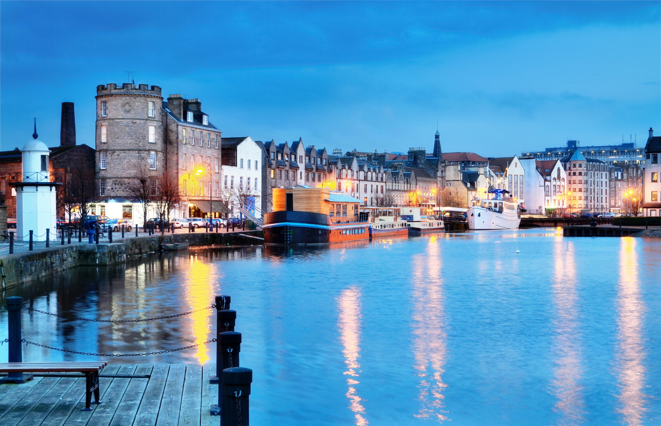 A scenic view of a harbor at dusk, featuring boats docked along the waterfront. The sky is a mix of blue and gray hues, while lights from buildings reflect on the water, creating a serene atmosphere. The architecture is a blend of modern and historic styles.