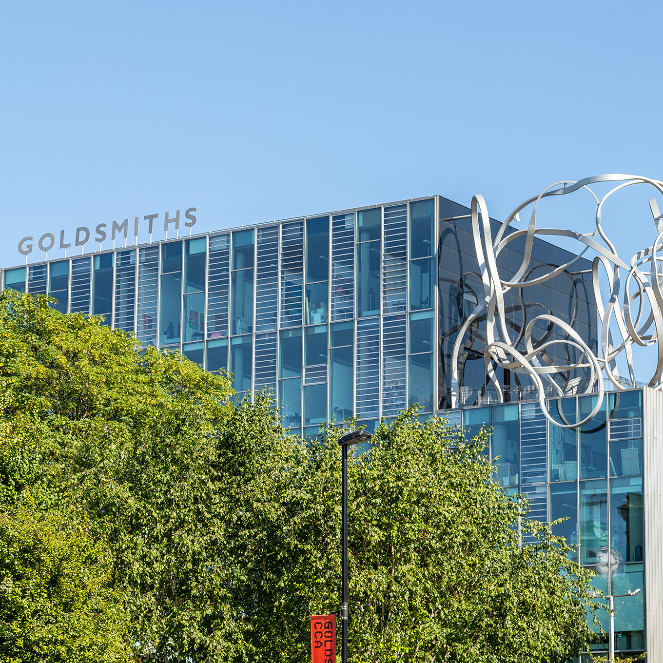 Modern glass building with "GOLDSMITHS" signage on the roof, surrounded by trees. A silver abstract sculpture is visible in front of the building against a clear blue sky.