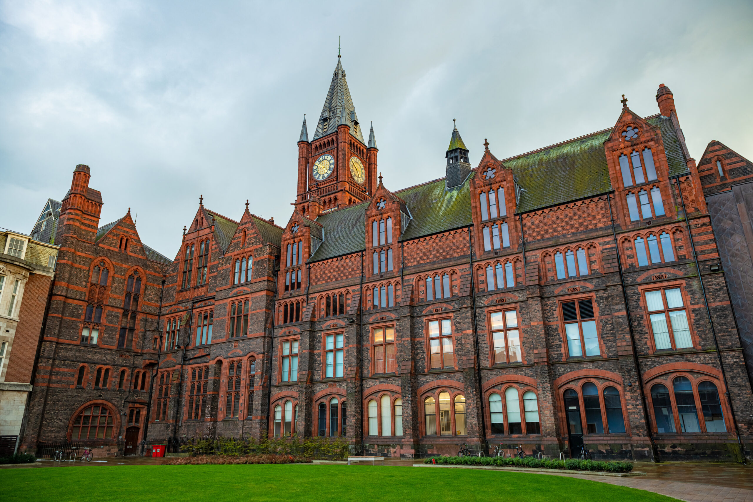 Historic red-brick building with a clock tower, featuring Gothic architectural elements like arched windows and spires, set against a cloudy sky. A manicured green lawn stretches across the foreground.