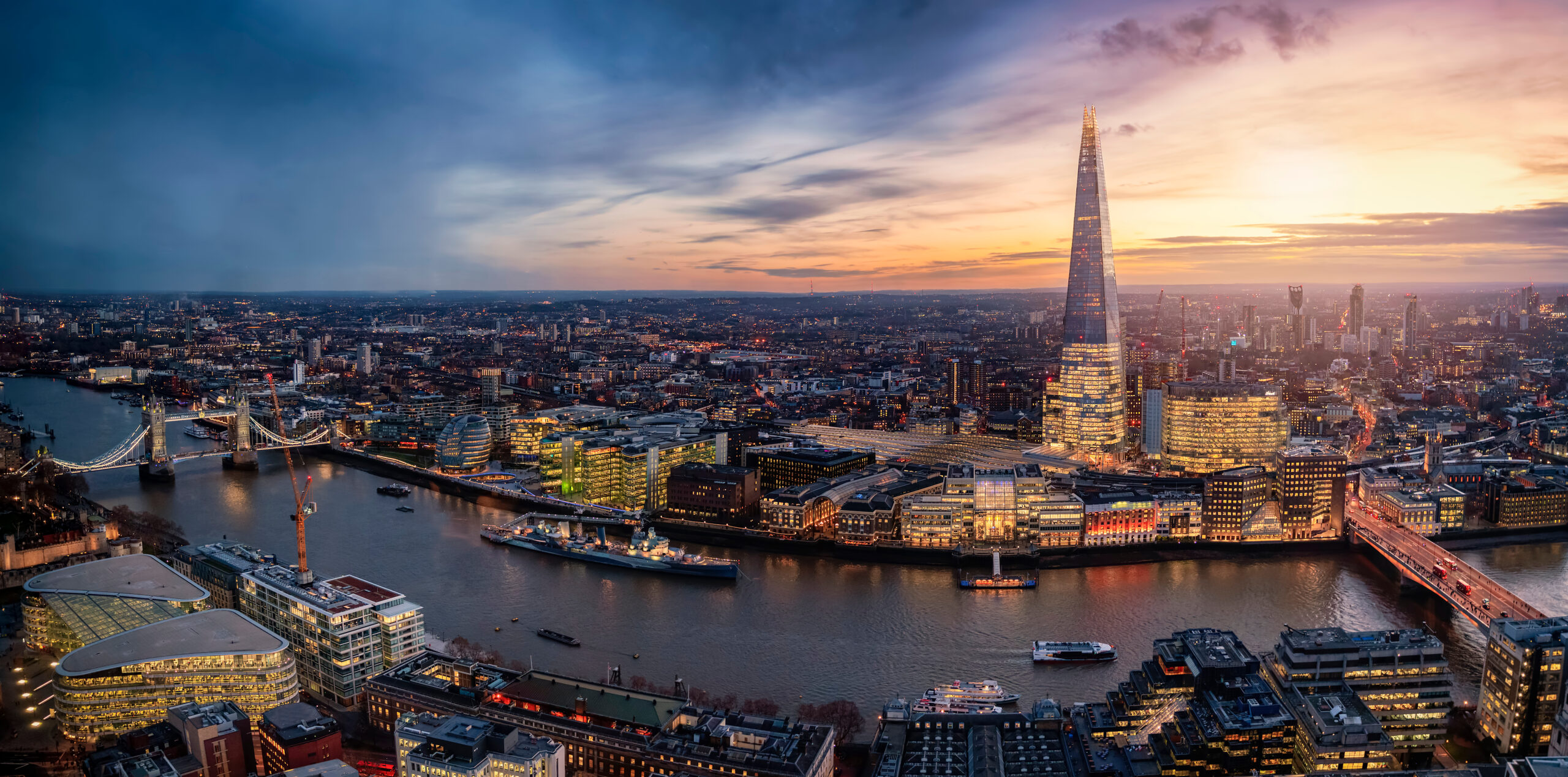 Aerial view of London at dusk, featuring the illuminated Shard skyscraper and Tower Bridge over the River Thames. The city skyline is visible against a colorful sunset. Buildings are lit, reflecting in the water below.