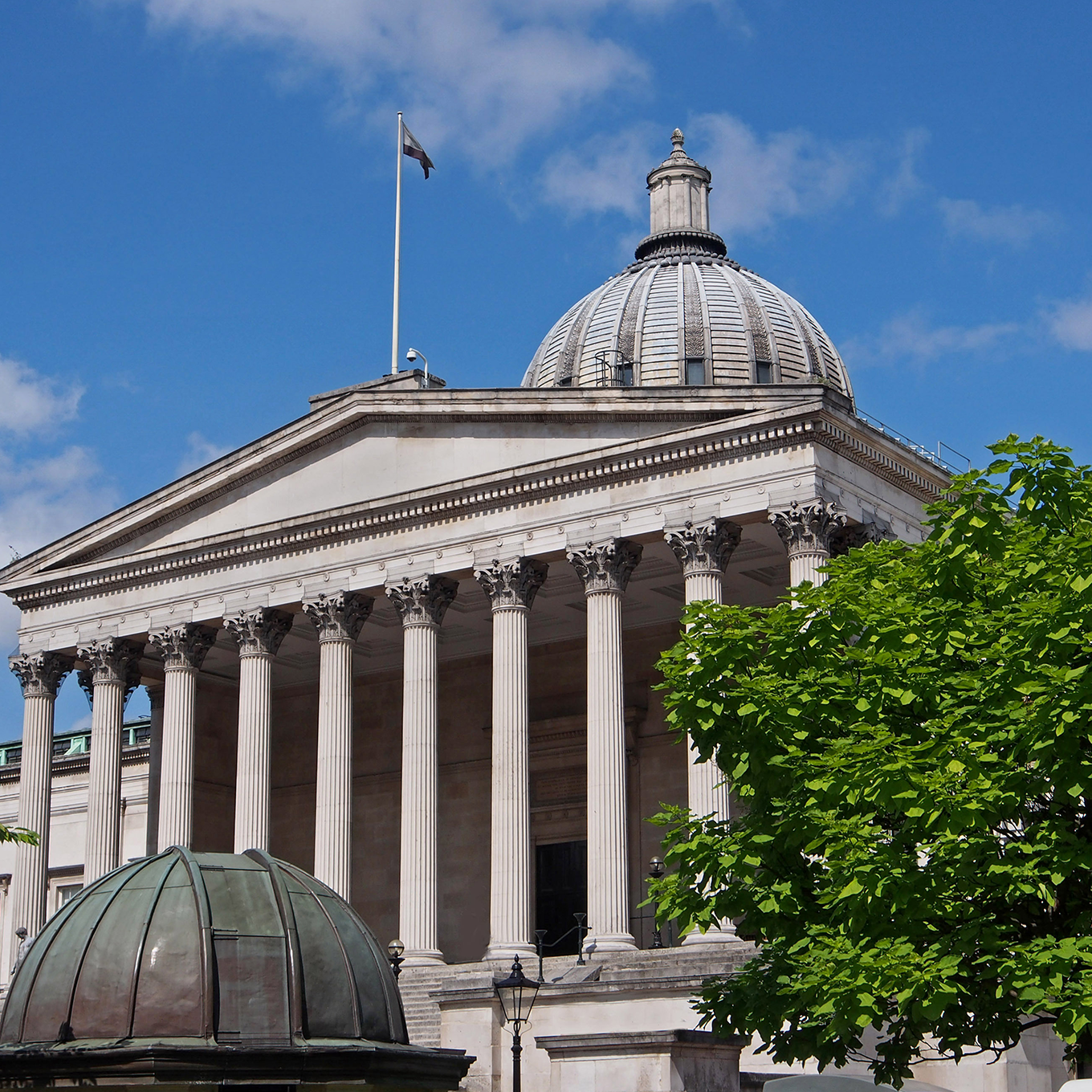A neoclassical building with a grand entrance featuring eight large columns and a prominent dome on top. The structure is white and stands against a blue sky, with a leafy green tree partially in view on the right.