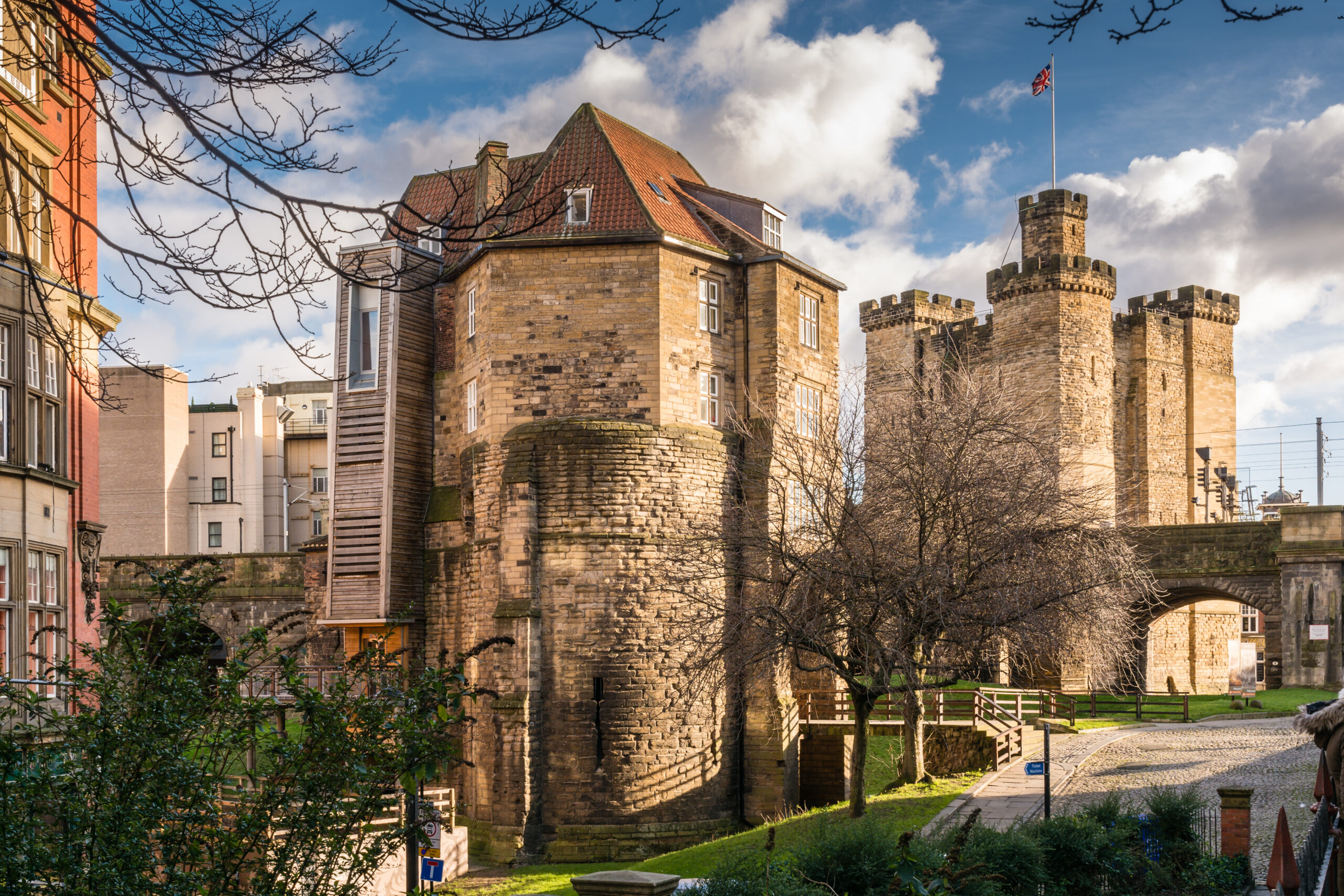 Historic stone castle with two large towers and a flag flying in the background. Leafless trees and brick path in the foreground, with a partly cloudy blue sky above. Surrounding buildings are visible, accentuating the old architecture.