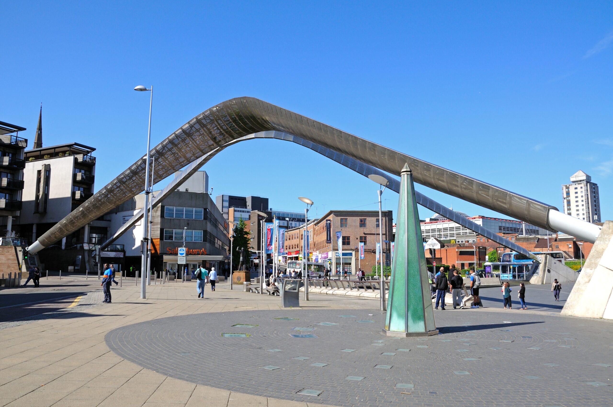 A modern architectural sculpture arches across the street in a public plaza near Coventry's student accommodation. People walk and interact beneath the structure, with surrounding buildings, including shops and residential areas, visible under a clear blue sky.