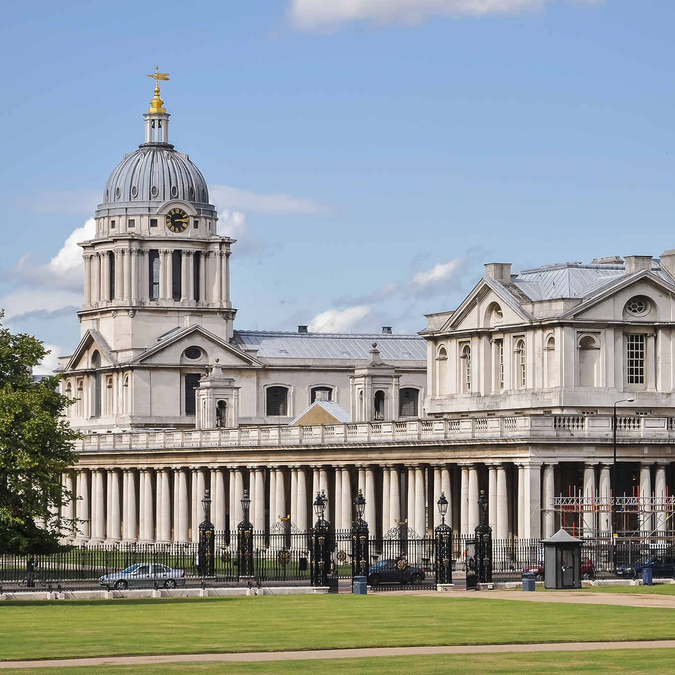 Historic European building with a prominent dome, classical columns, and intricate architectural details under a clear blue sky. Green lawn and wrought iron fencing in the foreground.