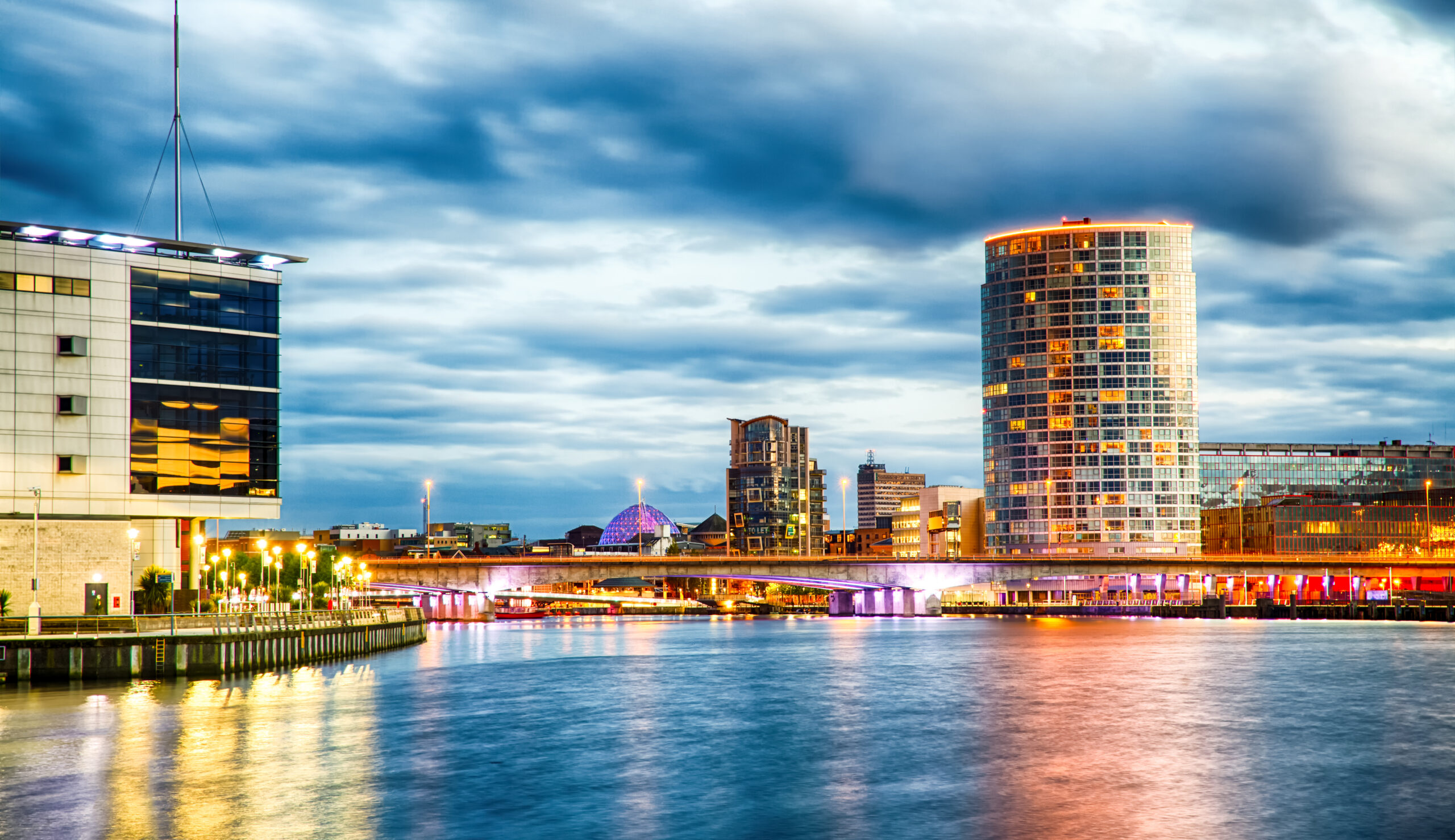 A vibrant cityscape at dusk featuring a tall, cylindrical building with glowing windows, a modern bridge lit with purple lights, and an expansive waterfront reflecting the colorful illumination under a cloudy sky.