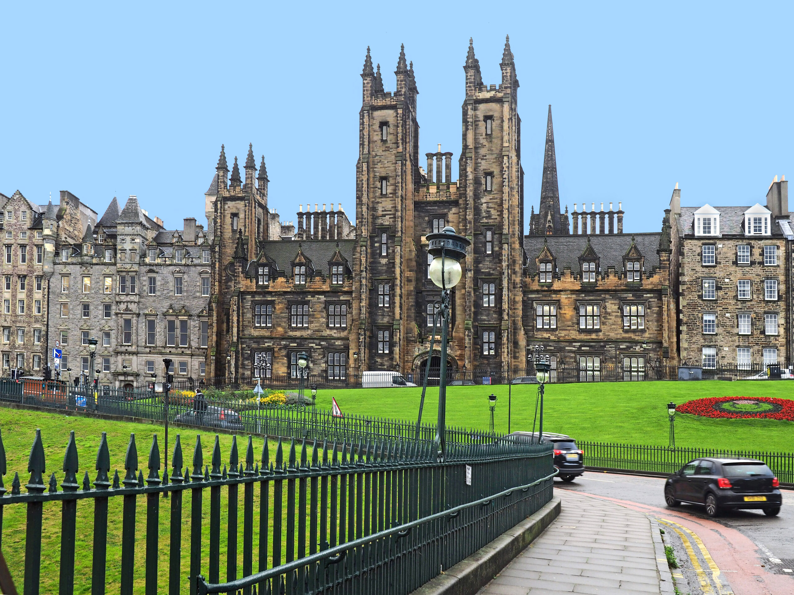 Gothic-style building with tall spires, surrounded by green lawns and a black iron fence. Two cars drive on a curving road in the foreground. Clear blue sky in the background.
