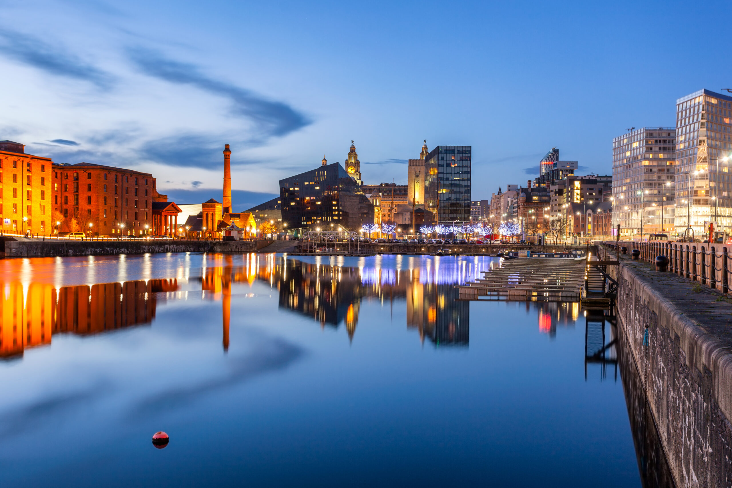 A serene night view of a waterfront cityscape. Buildings with illuminated windows and warm lights reflect on the calm water. The sky is a deep blue with few clouds, and a buoy floats in the foreground.