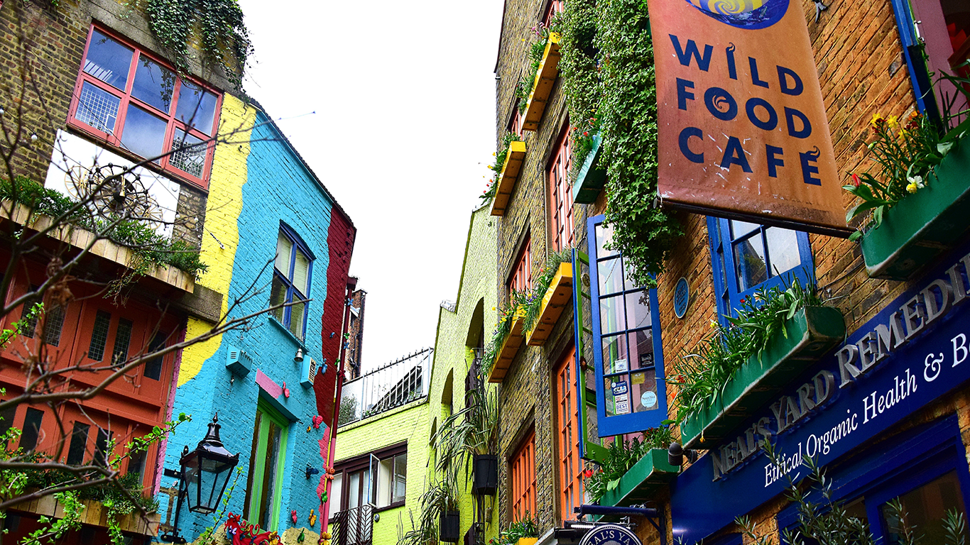 Colorful buildings in a narrow alleyway, adorned with plants and flowers. A sign reads "Wild Food Café." The vibrant walls are painted in blue, yellow, red, and green, creating a lively and artistic atmosphere.