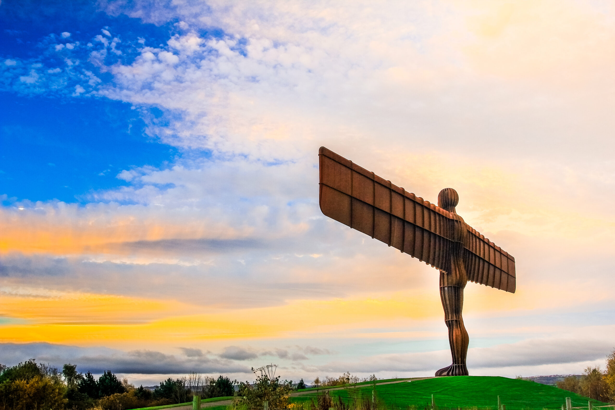 The Angel of the North sculpture stands on a grassy hill under a vibrant sky with orange, yellow, and blue hues during sunset. The large metal angel has outstretched wings and is silhouetted against the colorful backdrop.