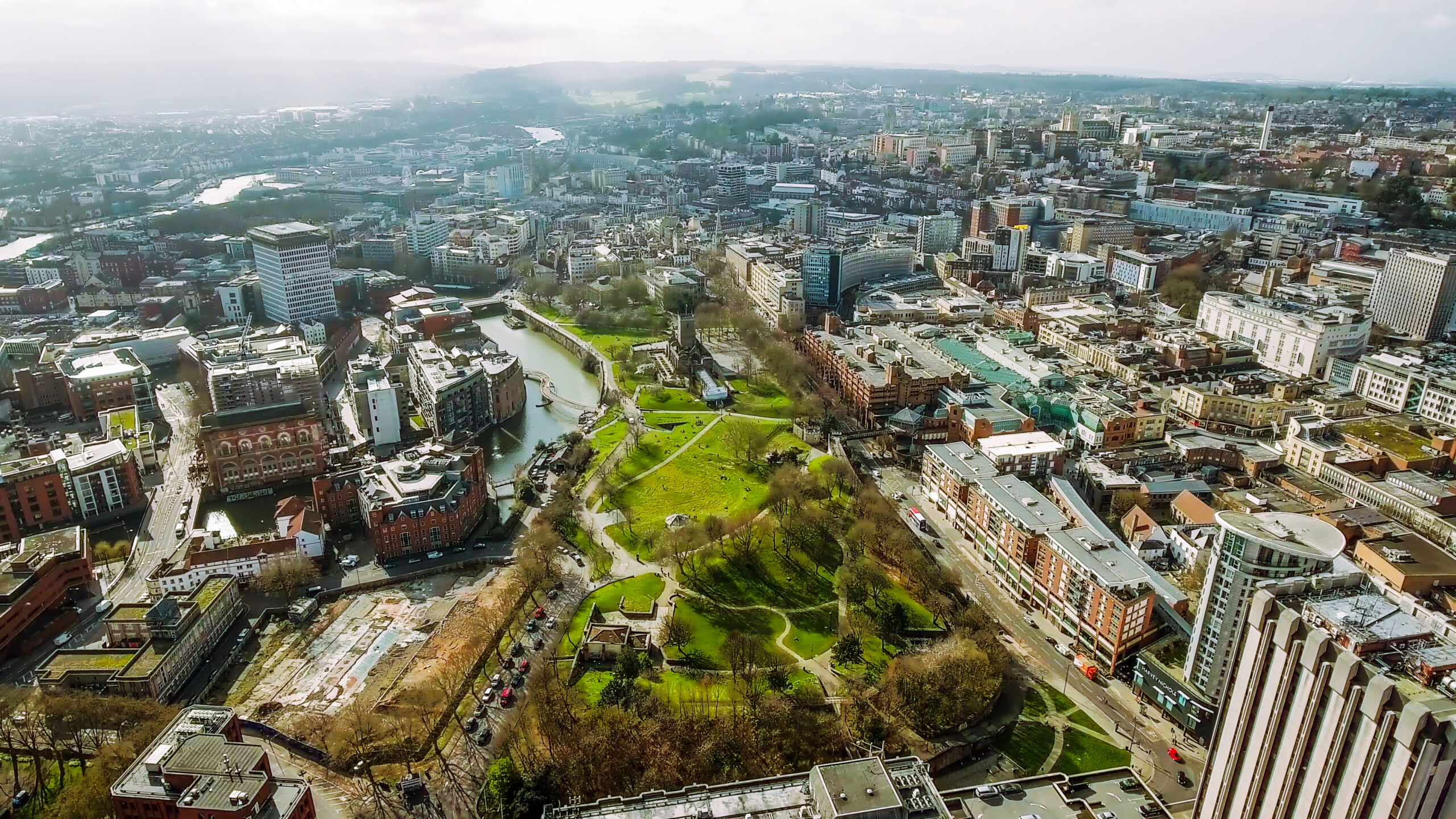 Aerial view of a city with a large green park at the center, surrounded by numerous buildings. A river winds through the cityscape, and roads are visible. The sky is overcast, casting a soft light over the urban landscape.