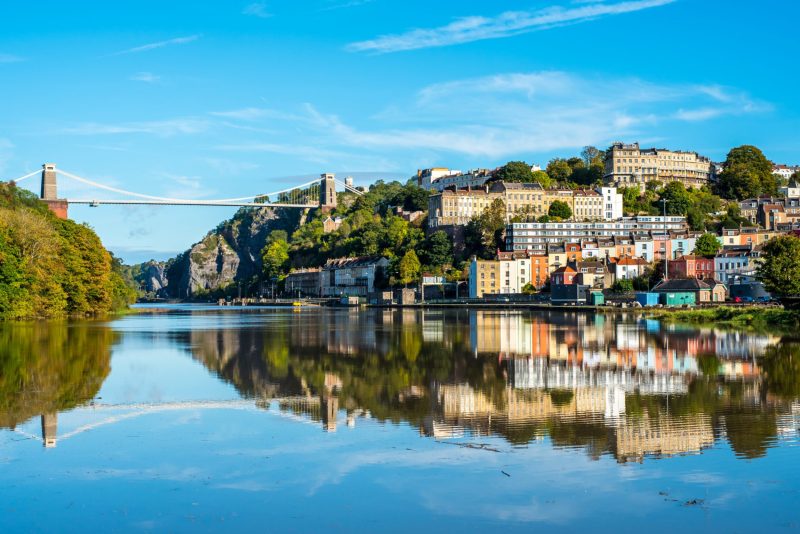 The image shows a scenic view of a river with the Clifton Suspension Bridge in the distance. Colorful buildings line the riverbank, and the clear water reflects both the bridge and the surrounding landscape under a bright blue sky.