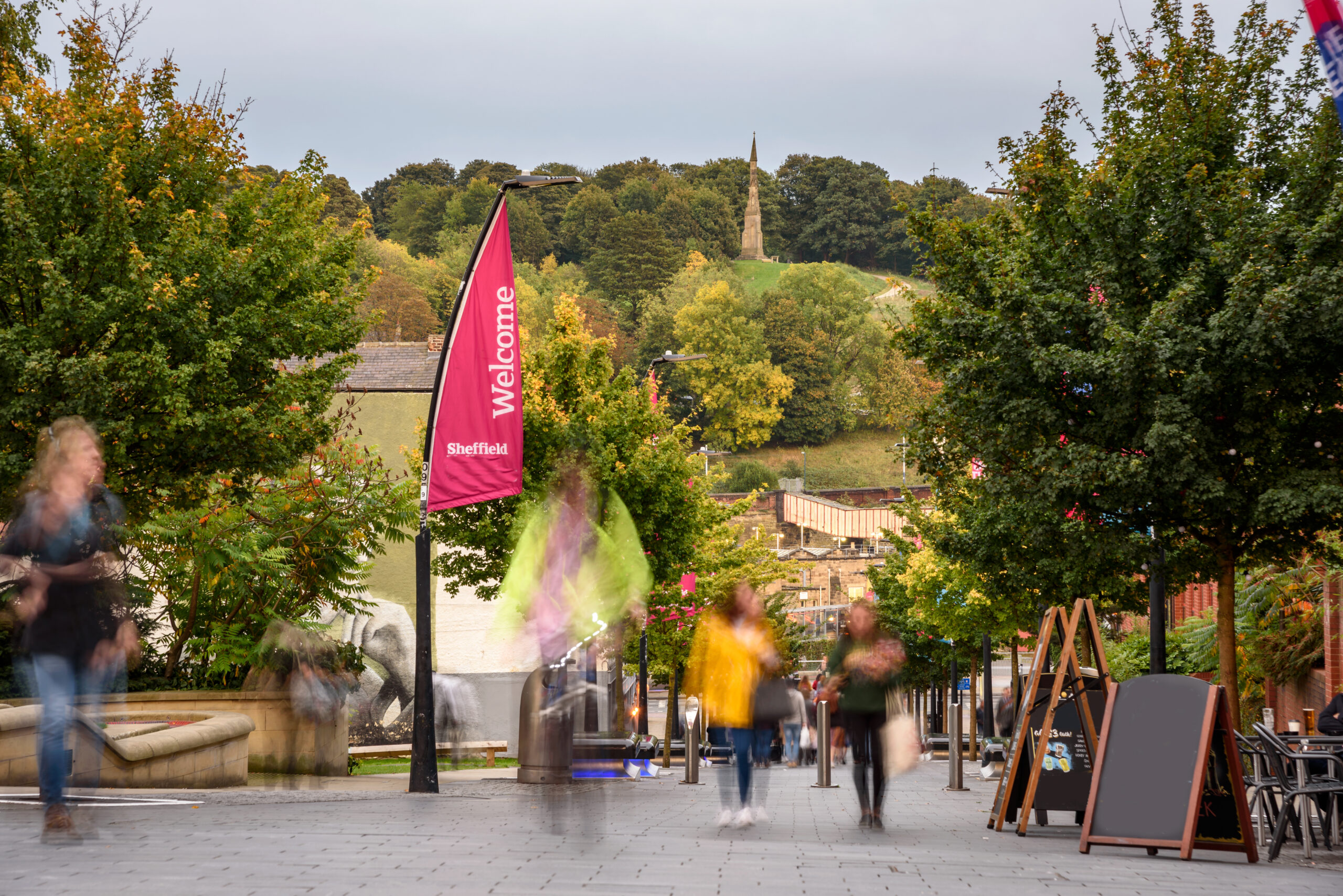 A bustling pedestrian walkway lined with trees, featuring a pink "Welcome to Sheffield" banner. People walk past, and in the distance, a hillside with a monument is visible. The scene captures an overcast day in an urban setting.