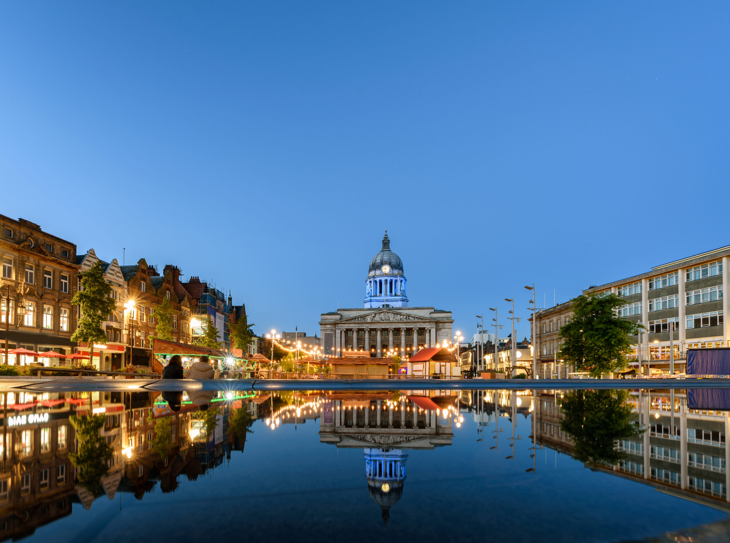 A grand neoclassical building with a large dome is beautifully illuminated in blue, reflected in a calm water surface at dusk. Surrounding it are lit buildings and trees on both sides, with a clear blue sky above.