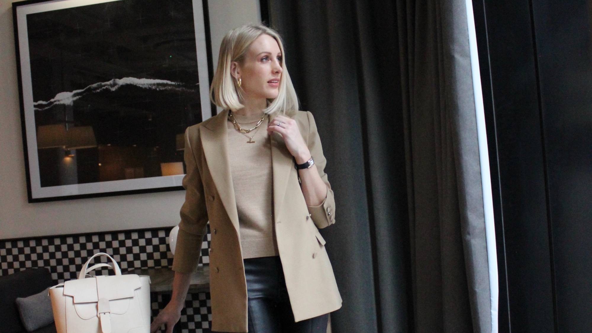 Charlotte Buttrick, in a beige blazer and black pants, stands by a window, embodying the perfect Dress For Interviews style. With her white bag and layered necklaces complementing the look, a framed abstract picture adds intrigue to her backdrop.