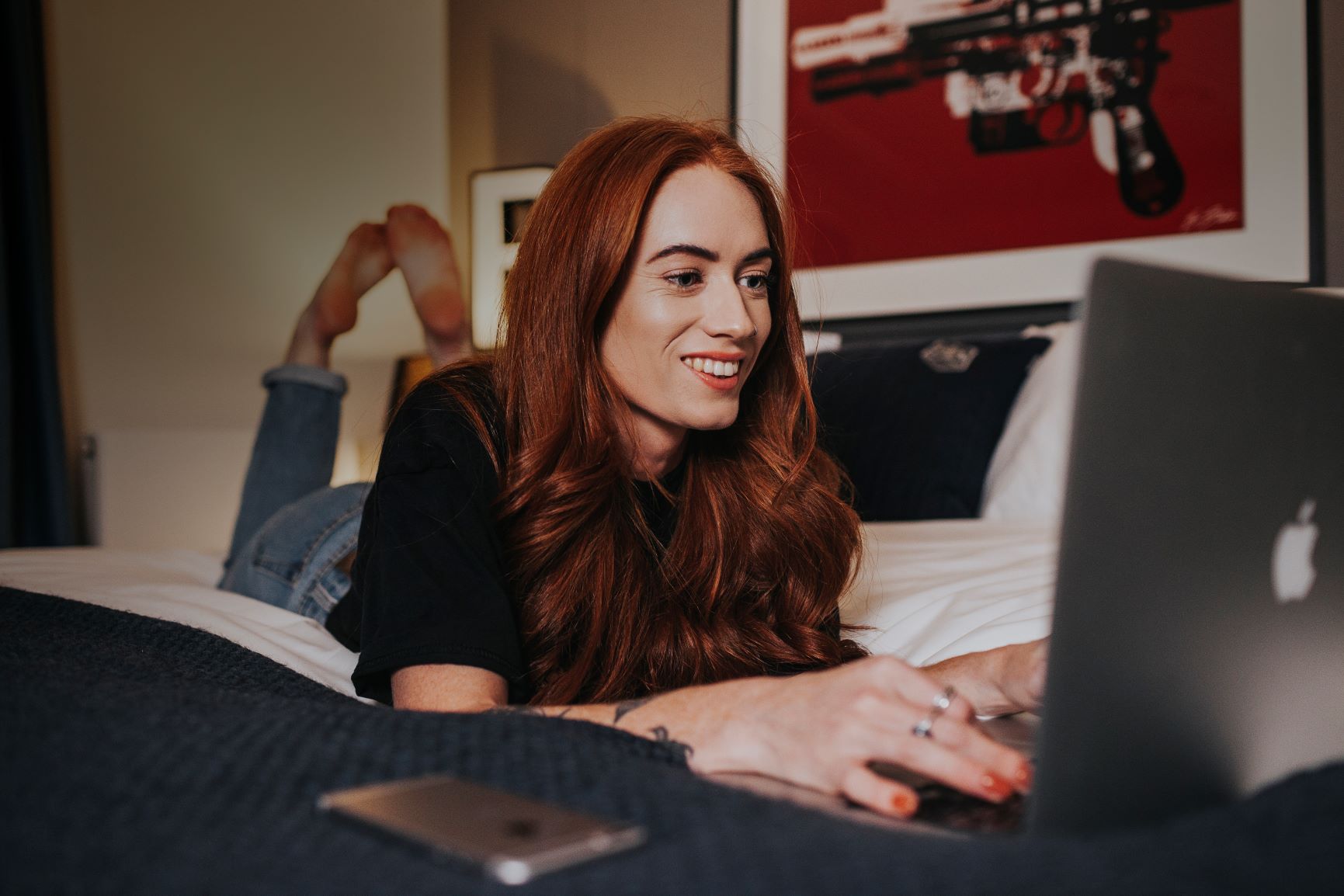 A woman with long red hair, wearing a black shirt and jeans, is staying motivated by working on her laptop while lying on a bed. A smartphone rests beside her as she takes creative steps; the background features artwork with a red and white theme.