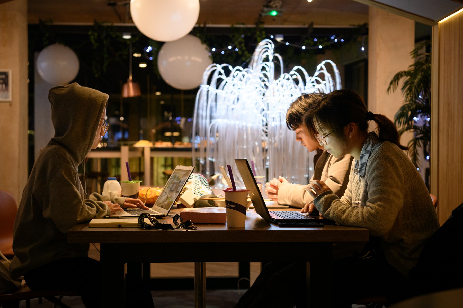 Three people sit at a table with laptops in a cozy indoor setting. A fountain with glowing lights is visible in the background. The room is warmly lit, creating a relaxed atmosphere.