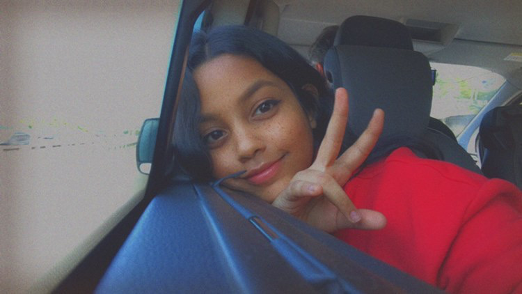 A young woman with long dark hair smiles and flashes a peace sign while leaning on a car window. Wearing a red top, Arya from Vita Student exudes joy as the colorful scenery of India blurs by outside the vehicle.