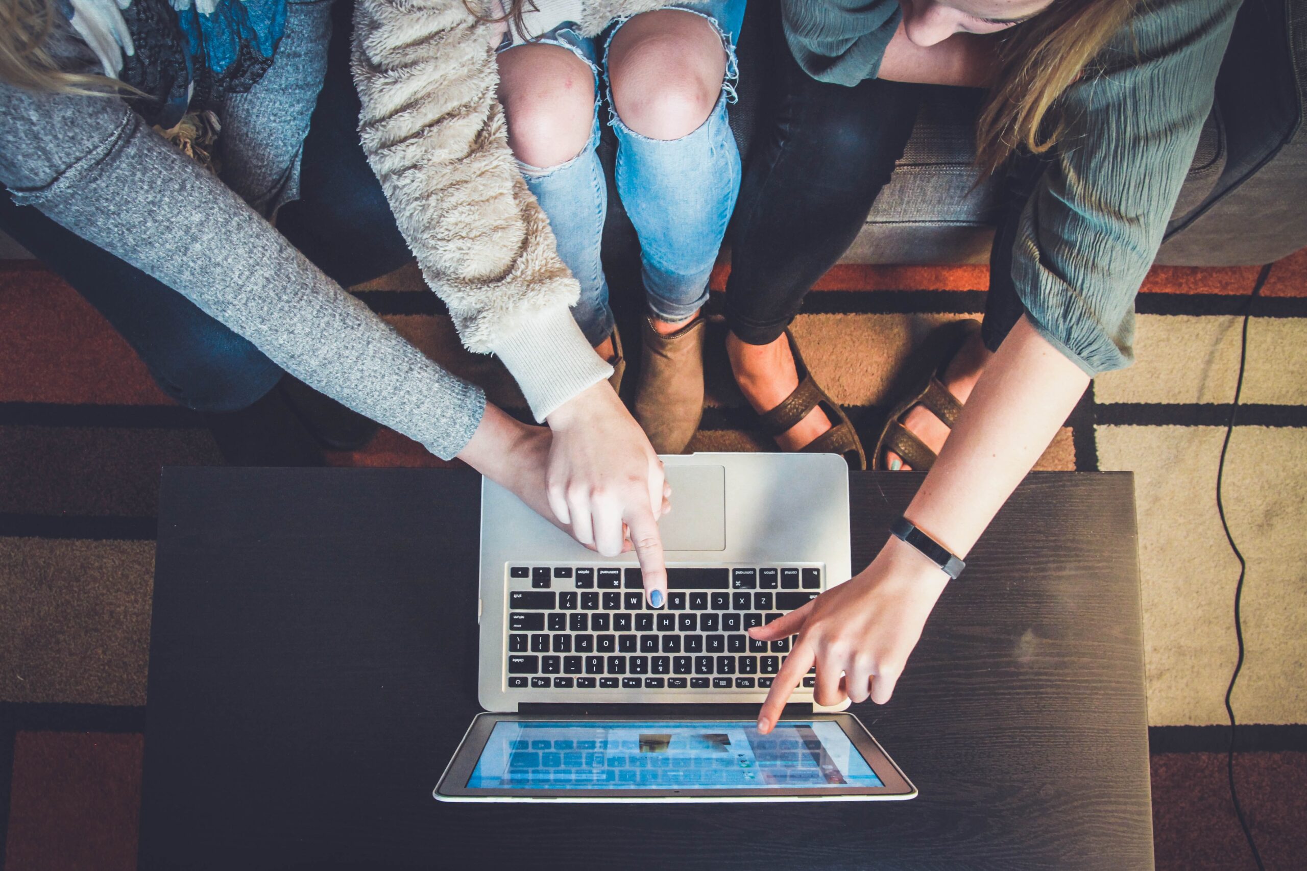 Three people, perhaps part of a student startup, are sitting on a couch, pointing and looking at a laptop screen on the table. Viewed from above, their hands and the bright display suggest they are collaborating intensely or discussing content to ace their next big idea.