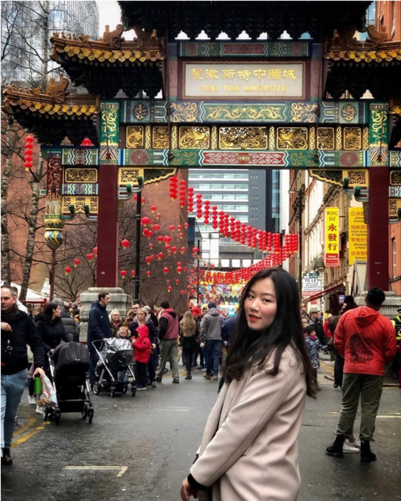 A woman stands in a bustling Chinatown street adorned with hanging red lanterns. Behind her, a traditional Chinese archway is visible. The area is crowded with people, some pushing strollers, enjoying the festive atmosphere.