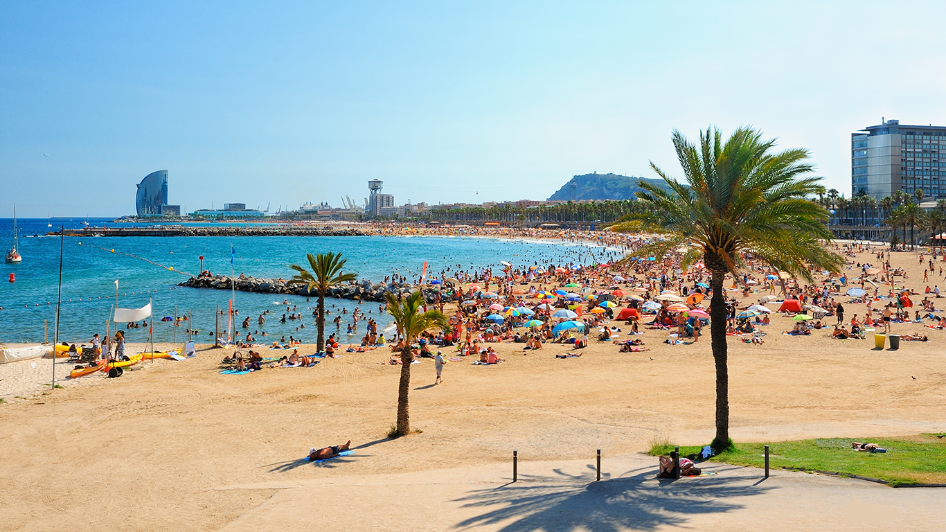 Sunny beach scene reminiscent of Barcelona, with a crowded shoreline. People are sunbathing, swimming, and walking among colorful umbrellas scattered across the sand. The blue sea glistens on the left, with palm trees and city buildings in the background under a clear sky.
