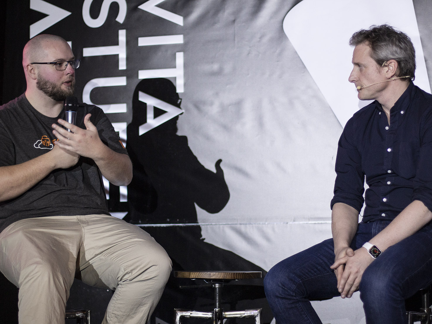 Two men are seated on stools having a conversation. The man on the left wears a dark shirt and gestures with his hands, holding a cup. The man on the right, sporting a blue shirt and headset microphone, seems engaged. A large, partially obscured "Tech Edition" sign looms behind them, hinting at Big Talks ahead.