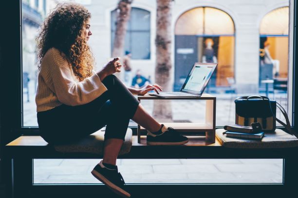 A person with curly hair is sitting on a windowsill, crafting compelling CV tips on a laptop. They're wearing a sweater, jeans, and sneakers. A backpack and notebook are beside them. The window shows an outdoor scene with blurred pedestrians.