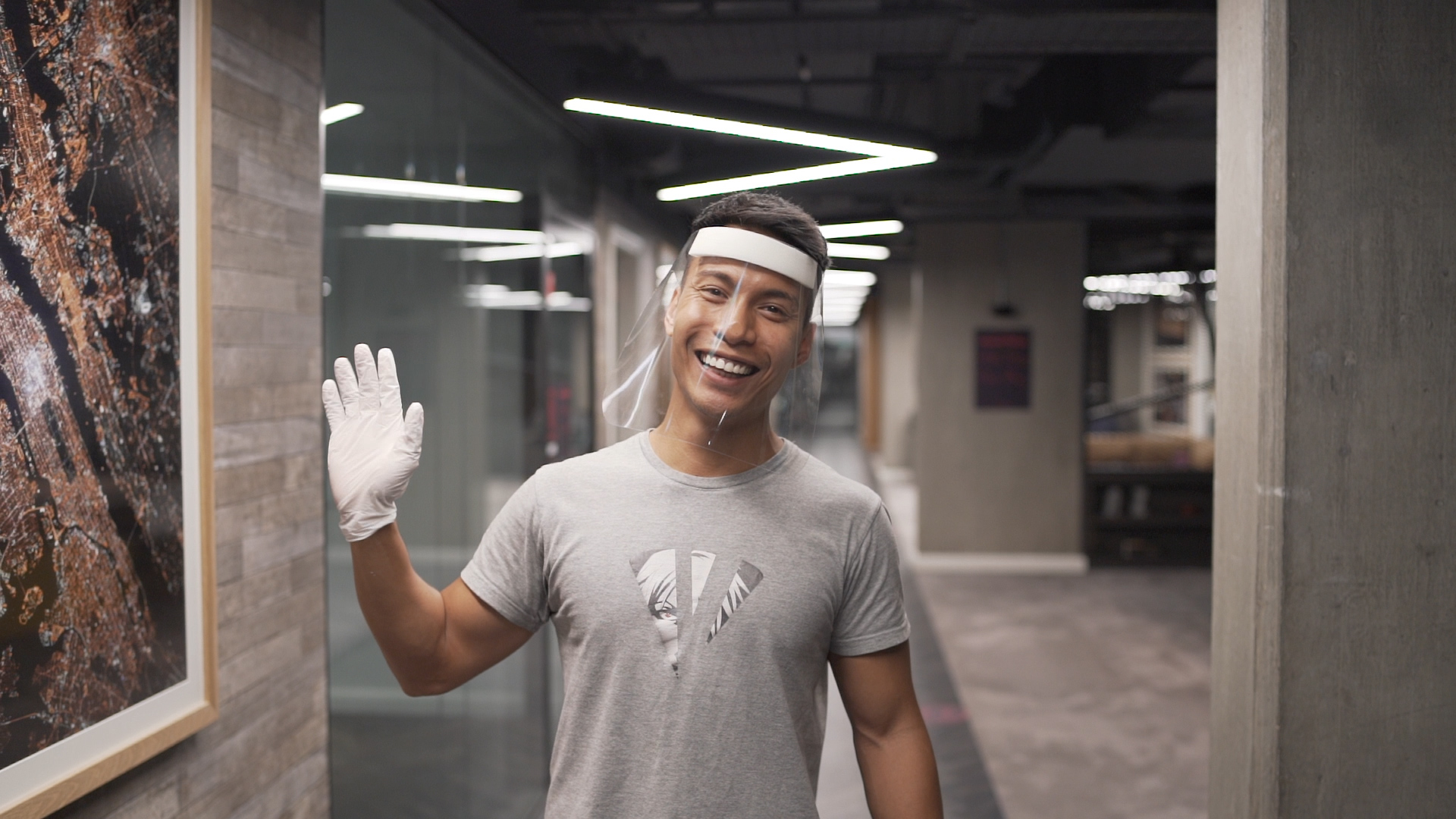 A person wearing a face shield, gloves, and a gray t-shirt waves and smiles in a hallway with modern decor and bright overhead lighting.