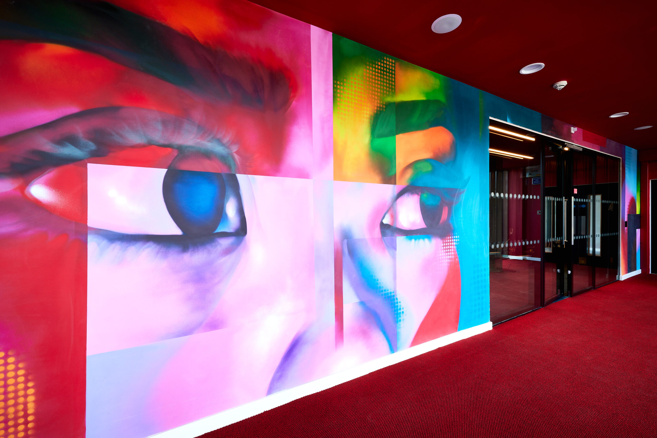 A vibrant, colorful mural by graffiti artist HazardOne depicts close-up eyes on a wall. The artwork uses bold hues of pink, blue, yellow, and green. The mural is adjacent to a glass door with a red carpeted floor leading into the room.