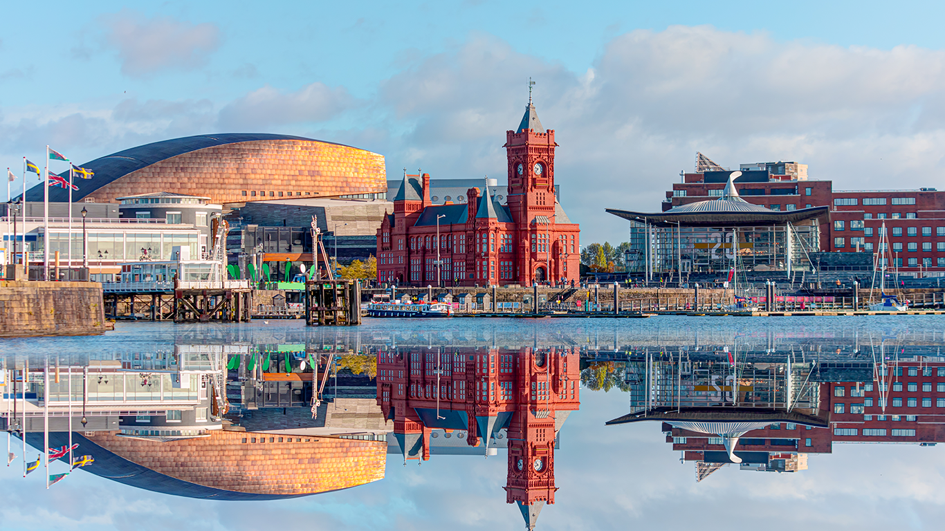 Experience a stunning waterfront view of Cardiff Bay as you explore things to do near the iconic Wales Millennium Centre, the historic red-brick Pierhead Building, and the modern Senedd building, all beautifully reflected in the calm water under a picturesque blue sky with clouds.