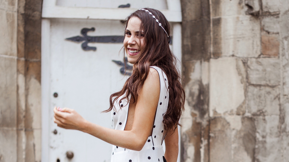 Ciandra, a woman with long brown hair and a headband, smiles near a weathered stone wall and a white door. She is wearing a sleeveless white dress adorned with black polka dots, exuding an air of confident insight.
