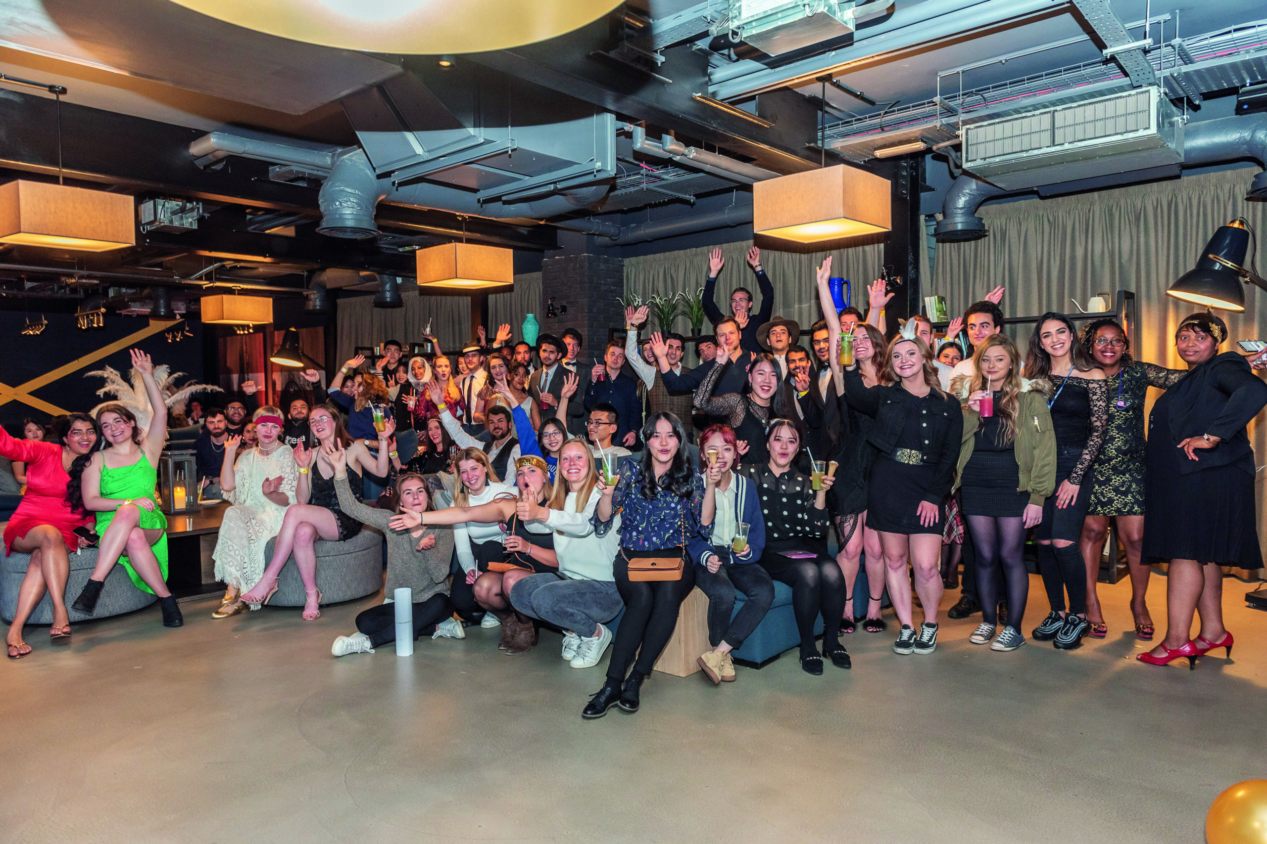 A large group of people pose enthusiastically in a stylish indoor setting with modern UK decor. Some individuals are dressed in vibrant, colorful attire, while others wear casual outfits. The atmosphere is lively and joyful, capturing the essence of English conviviality.