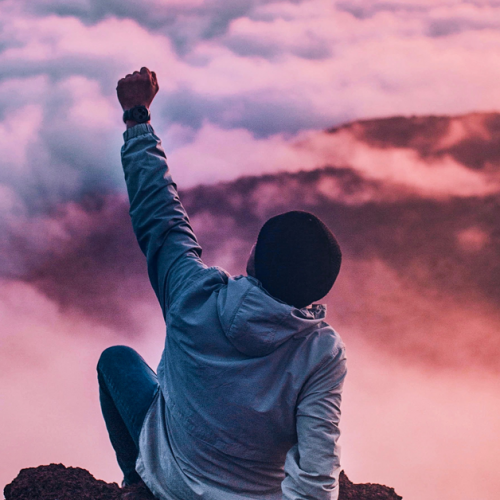 A person in a hoodie and jeans sits on a rocky cliff, raising one arm triumphantly, casting away the freshers' fear. They are surrounded by clouds with a pink and purple sky, suggesting a sunrise or sunset.