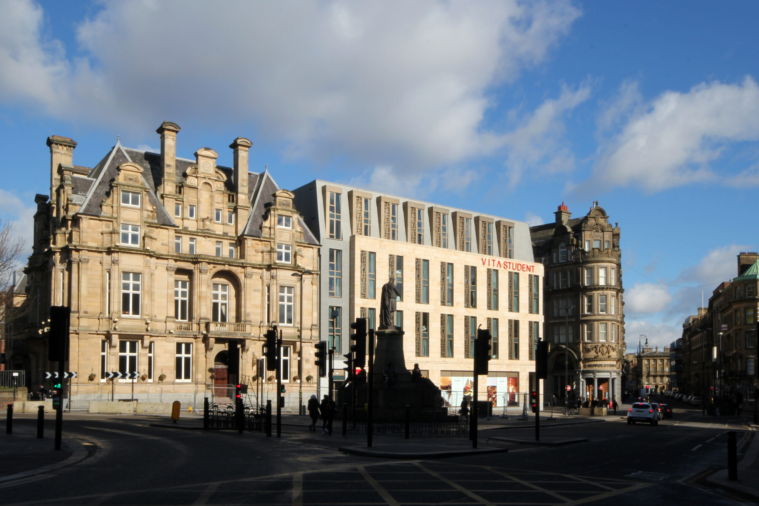 Street view of a cityscape featuring historic and modern buildings. The foreground shows a roundabout with a statue. The sky is partly cloudy, and cars are at traffic lights.